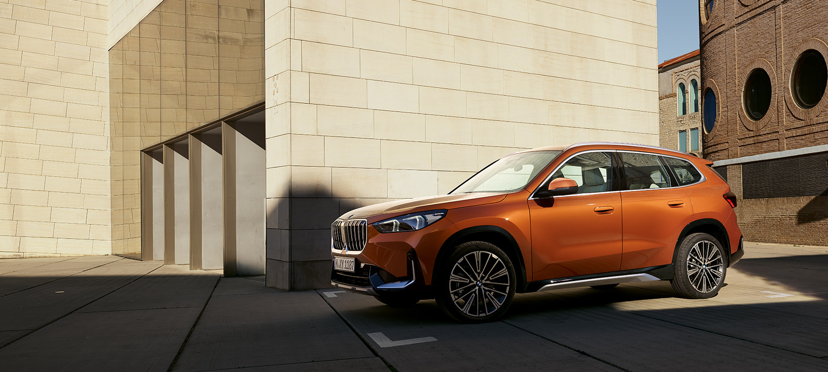 BMW U11 X1 xLine Utah orange metallic vue de trois quarts avant à l’arrêt devant un bâtiment